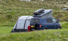 Das Bild zeigt einen VW Bus stehend von schräg hinten rechts fotografiert. An der linken Fahrzeugseite (im Bild links) ist ein Busvorzelt angebracht.