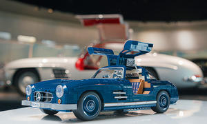 Das Bild zeigt den Mattel Brick Shop Mercedes 300 SL von schräg vorne links mit geöffneten Flügeltüren. Im Hintergrund ist ein echter Mercedes 300 SL zu sehen.