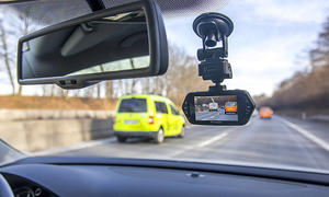 Das Bild zeigt eine Dashcam, welche per Saugnapf an der Innenseite einer Windschutzscheibe in einem Auto rechts des Innenspiegels montiert ist. Die Kamera filmt den Verkehr auf einer Autobahn vor dem Fahrzeug in dem sich die Kamera befindet.