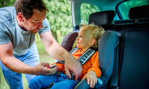 Das Bild zeigt eine Person, die durch die geöffnete Hintertüre auf der rechten Fahrzeugseite ein Kind in einem Kindersitz anschnallt. Ansicht vom Fahrersitz aus.