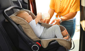 Das Bild zeigt ein Kleinkind in einem vorwärtsgerichteten Reboarder auf der Rückbank eines Autos. Das Kind hält eine Babyflasche in der Hand, eine Person steht an der geöffneten Schiebetüre des Autos und schnallt das Kind an.
