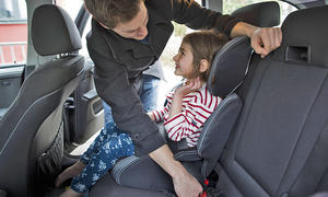 Ein Kind wird von einer erwachsenen Person auf einer Kindersitzerhöhung mit Rückenlehne im Auto auf der Rücksitzbank angeschnallt.