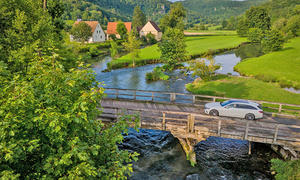 Das Mercedes E 220 d 4Matic T-Modell fährt über eine Holzbrücke. Im Hintergrund eine idyllische Landschaft und Häuser.