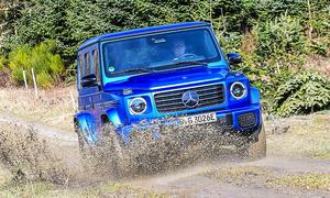 Blick von schräg vorne auf den fahrenden Mercedes G 580 EQ, der gerade durch eine tiefe Matschpfütze fährt.