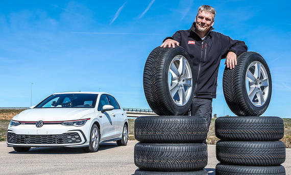 Der "All Zeason"-Allwetterreifen der AUTO ZEITUNG - Daniela Loof Redakteur Martin Urbanke posiert stolz hinter einem Stapel Reifen, im Hintergrund steht der Testwagen VW Golf GTI.