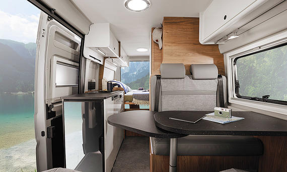 Corigon Campervan (2025) - Corigon Das Bild zeigt den Wohnraum des Corigon Campervan (2025). Das Foto wurde im Cockpit mit Blick nach hinten aufgenommen und zeigt die Sitzgruppe, die Küche und das Heckbett.