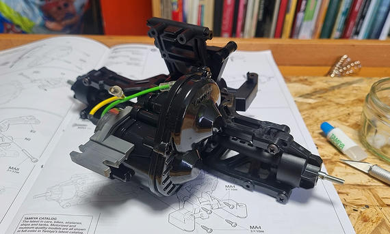 Das Bild zeigt das RC-Auto Tamiya DT-04 Fighter Nxgen im Aufbau. Zu sehen ist die Hinterachs-Konstruktion samt Antrieb auf der aufgeklappten Anleitung.