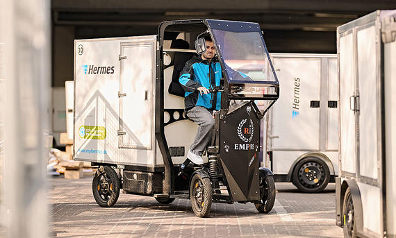 Das Bild zeigt ein Schwerlastenrad des Paketzustelldienstes Hermes. Eine Person sitzt im Fahrerhaus des vierrädrigen Cargo Bikes.