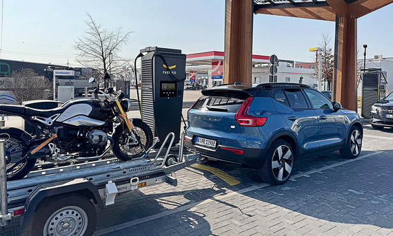 Der Volvo XC40 Recharge Extended Range mit Motorradanhänger an einer Ladestation, fotografiert von schräg hinten.