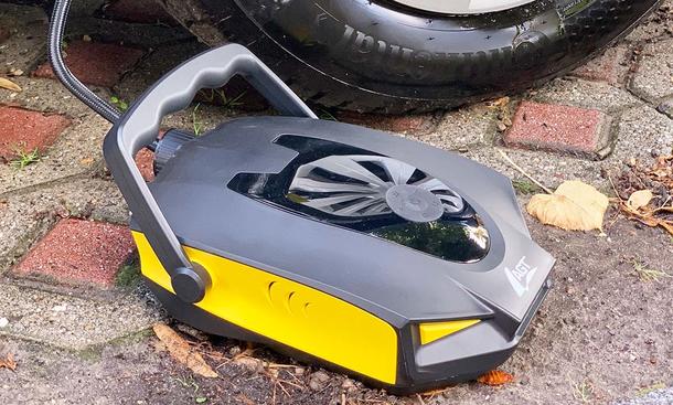 Akku-Luftpumpe Pearl AGT in der Einfahrt vor einem Autoreifen, fotografiert von schräg vorne.