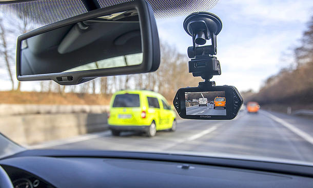 Das Bild zeigt eine Dashcam, welche per Saugnapf an der Innenseite einer Windschutzscheibe in einem Auto rechts des Innenspiegels montiert ist. Die Kamera filmt den Verkehr auf einer Autobahn vor dem Fahrzeug in dem sich die Kamera befindet.