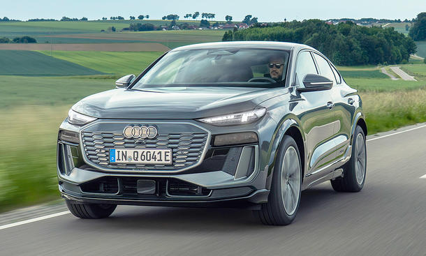 Der Audi Q6 Sportback e-tron (2025) fährt eine Landstraße im Grünen entlang, von vorne links fotografiert.
