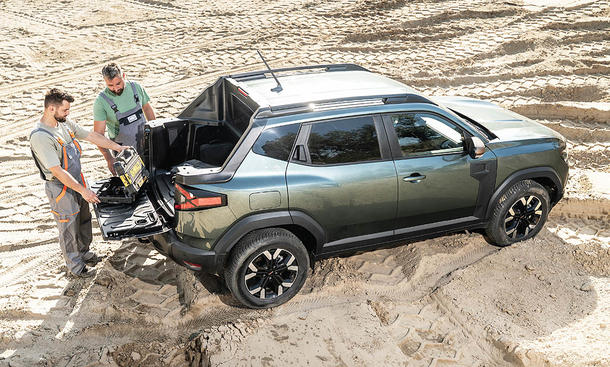 Der Dacia Duster Pick-up steht auf sandigem Untergrund, zwei Handwerker begutachten einen Werkzeugkoffer auf der Ladefläche.