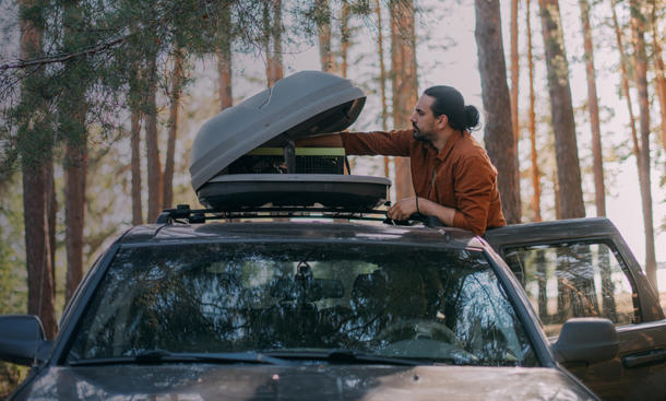 Mann öffnet Dachbox eines im Wald stehenden Autos.