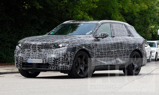 Ein Erlkönig des BMW iX5 M70 xDrive (2026) fährt auf einer Straße, von vorne links fotografiert.
