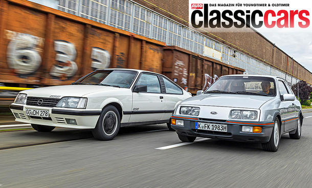 Opel Monza GSE und Ford Sierra XR4i fahren nebeneinander durch ein Industriegebiet, von vorne fotografiert.