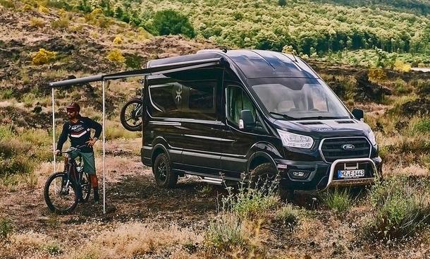 Die Außenaufnahme zeigt den Karmann Dexter 560 4x4 (2025) statisch von schräg vorne links. Vor dem Wohnmobil steht eine Person auf einem Mountainbike, die Markise ist ausgefahren.