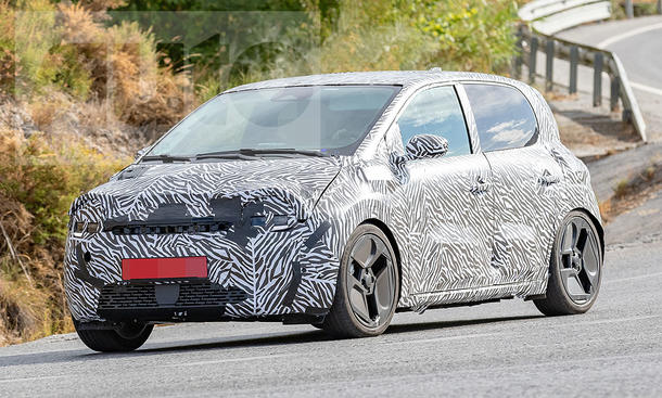 Der Renault Twingo E-Tech Electric (2026) fährt als Erlkönig auf einer Landstraße durch eine Rechtskurve, von vorne links fotografiert.