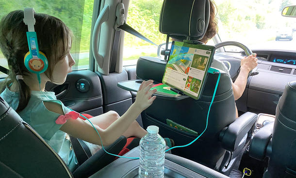 Mädchen auf der Rücksitzbank eines Autos beim Bedienen eines Tablets in einer Tablet-Halterung, fotografiert von hinten mit Blick auf Mädchen und Person am Steuer.