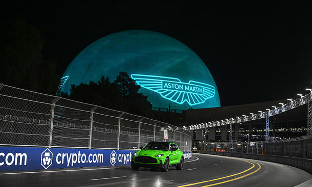 Der Aston Martin DBX auf einer leeren Straße fahrend, im Hintergrund sieht man groß das Markenlogo.