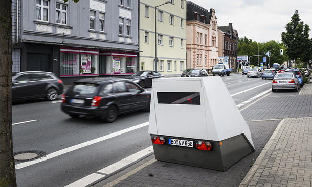Ein Blitzer-Anhänger an einem Straßenrand stehend, Fahrzeuge fahren daran vorbei.