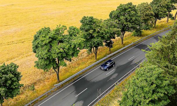 Blick von schräg vorne aus der Vogelperspektive auf den BMW i5 Touring, der über eine von Bäumen umgebene Landstraße fährt.