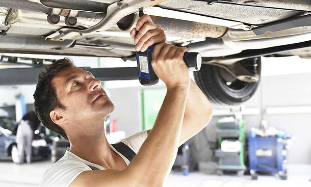 Ein Mann in Arbeitsmontur steht unter einer Hebebühne und arbeitet mit einer Bohrmaschine an einem Auto.