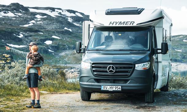 Die Außenaufnahme zeigt den Hymer Tramp S 585 (2025) in der Frontansicht vor einer Fjordlandschaft. Ein Mann mit Kind auf dem Arm steht daneben.