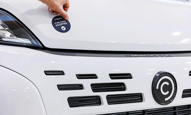 Das Bild zeigt die Motorhaube und den Kühlergrill des Corigon-Wohnmobil. Eine Hand ist in der oberen linken Bildecke zu sehen. Sie klebt einen Aufkleber mit der Aufschrift "Corigon Quality Control Accepted" auf die Motorhaube.