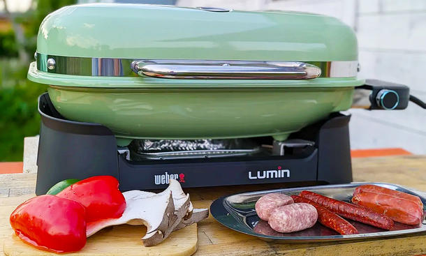 Der Weber Elektro-Tischgrill Lumin von vorne fotografiert. Auf dem Tisch ist zudem ein Tablett mit verschiedenen rohen Wurstsorten und ein Brett mit Paprika und Pilzen abgestellt.